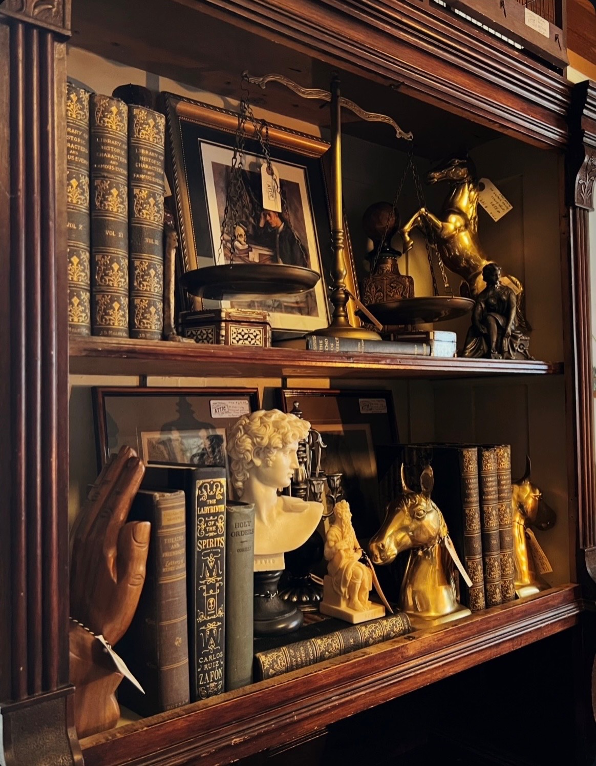 Antique bookshelf vignette with ornate books, brass horse sculpture, classical bust, balance scale, and framed artwork.